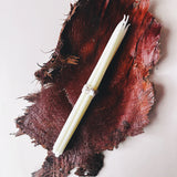 A bundle of white taper candles wrapped with a silver band, laid diagonally on a dark reddish-brown textured organic background against a light off-white backdrop.