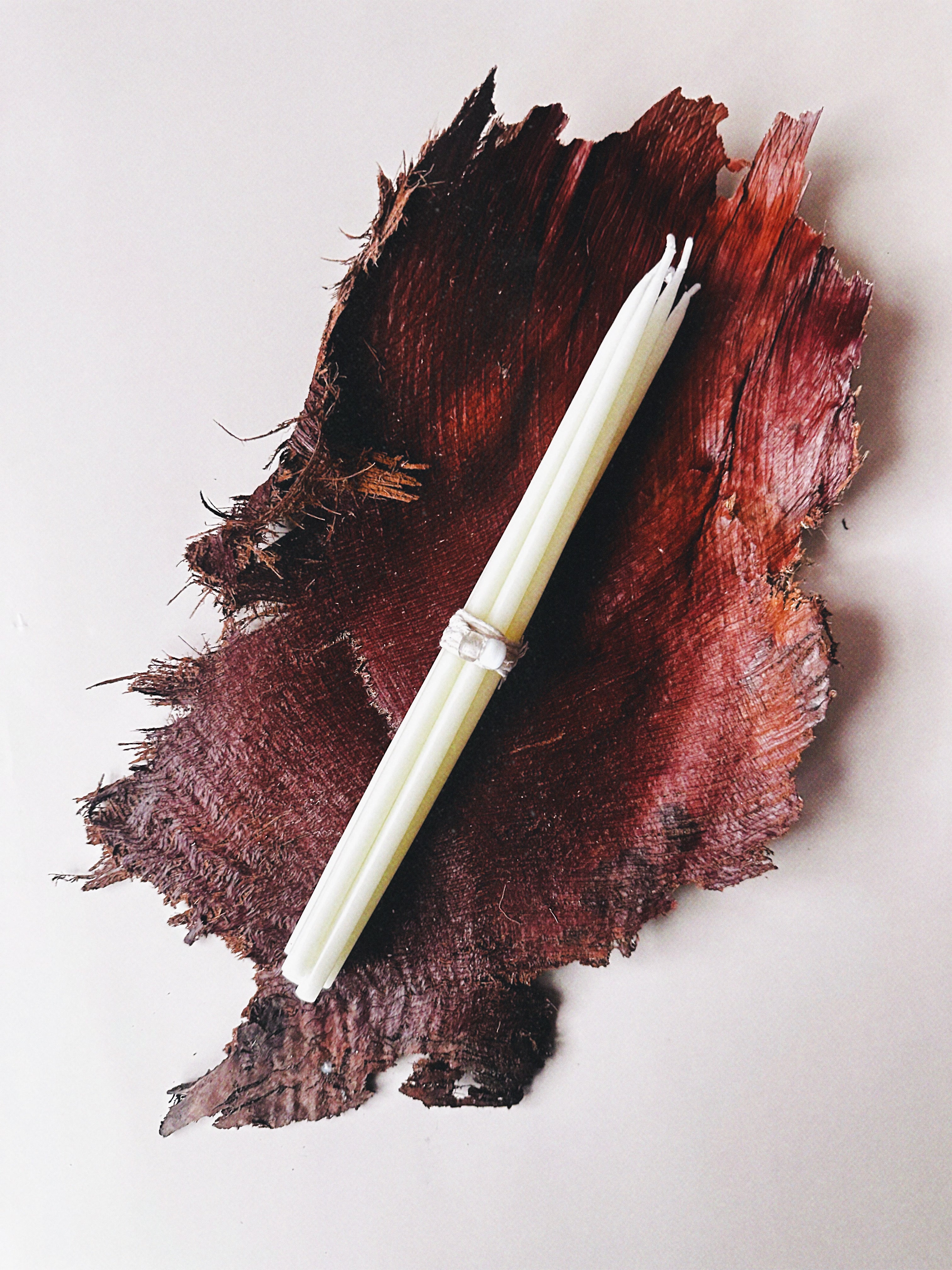 A bundle of white taper candles wrapped with a silver band, laid diagonally on a dark reddish-brown textured organic background against a light off-white backdrop.
