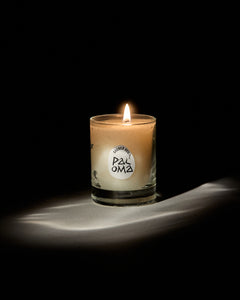 Paloma mini candle by San Maris lit and glowing on a draped white cloth against a black background