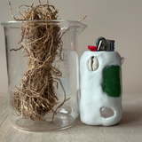Glass container with vetiver and a white ceramic lighter with green accents on a neutral background