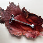 A pair of silver metal scissors with circular handles rests on a textured, dark red and brown piece of wood or leather against a plain off-white background.