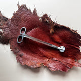 A pair of silver metal scissors with circular handles rests on a textured, dark red and brown piece of wood or leather against a plain off-white background.